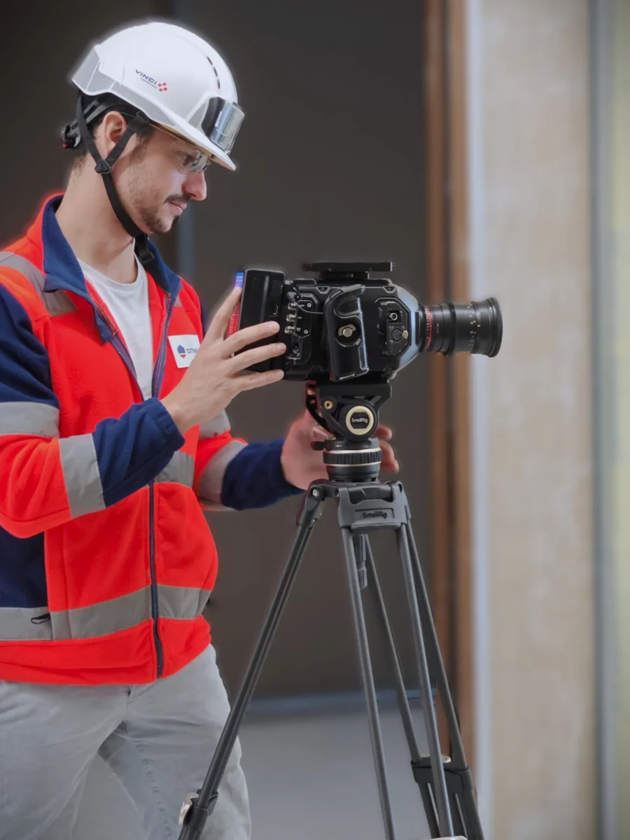 Axel Milon filmant sur un chantier, casque et gilet haute visibilité, caméra Blackmagic sur trépied